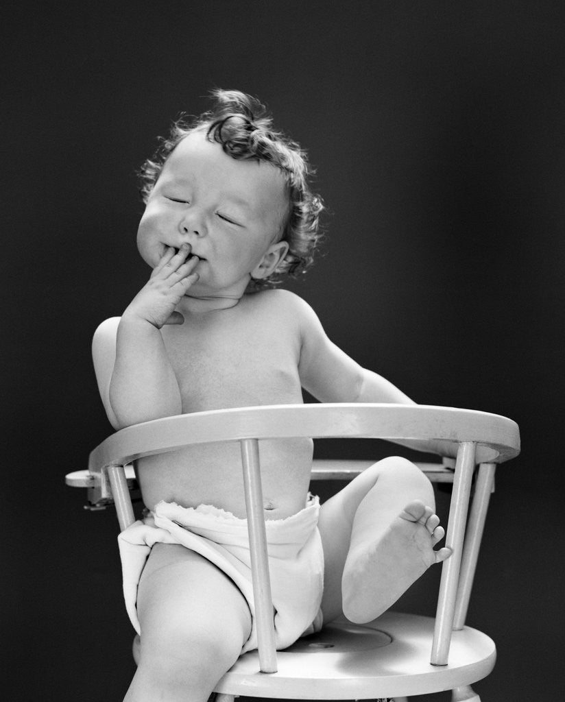 Detail of 1940s 1950s Baby Sitting Backwards In High Chair With One Leg In Air Eyes Closed and Hand On Mouth by Corbis