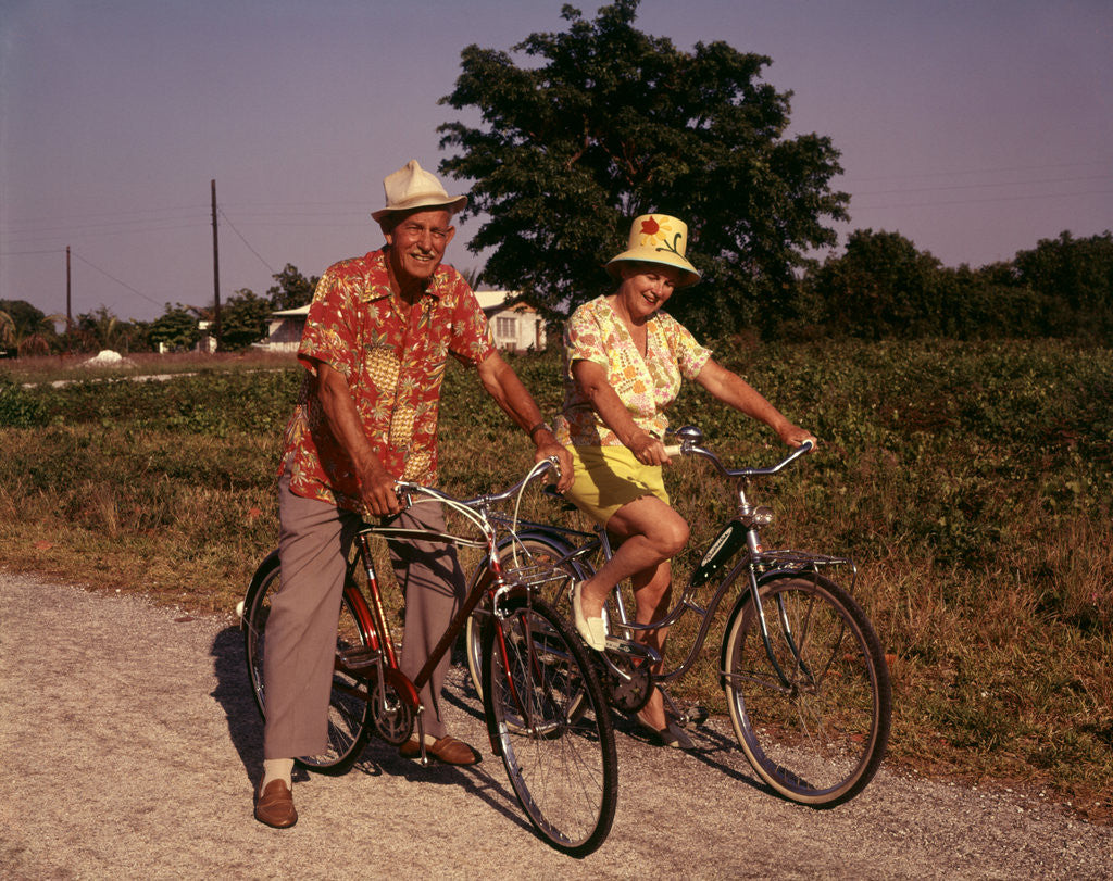 Detail of 1970s Senior Elderly Retired Couple Riding Bikes Wearing Straw Hats Hawaiian Print Shirts by Corbis