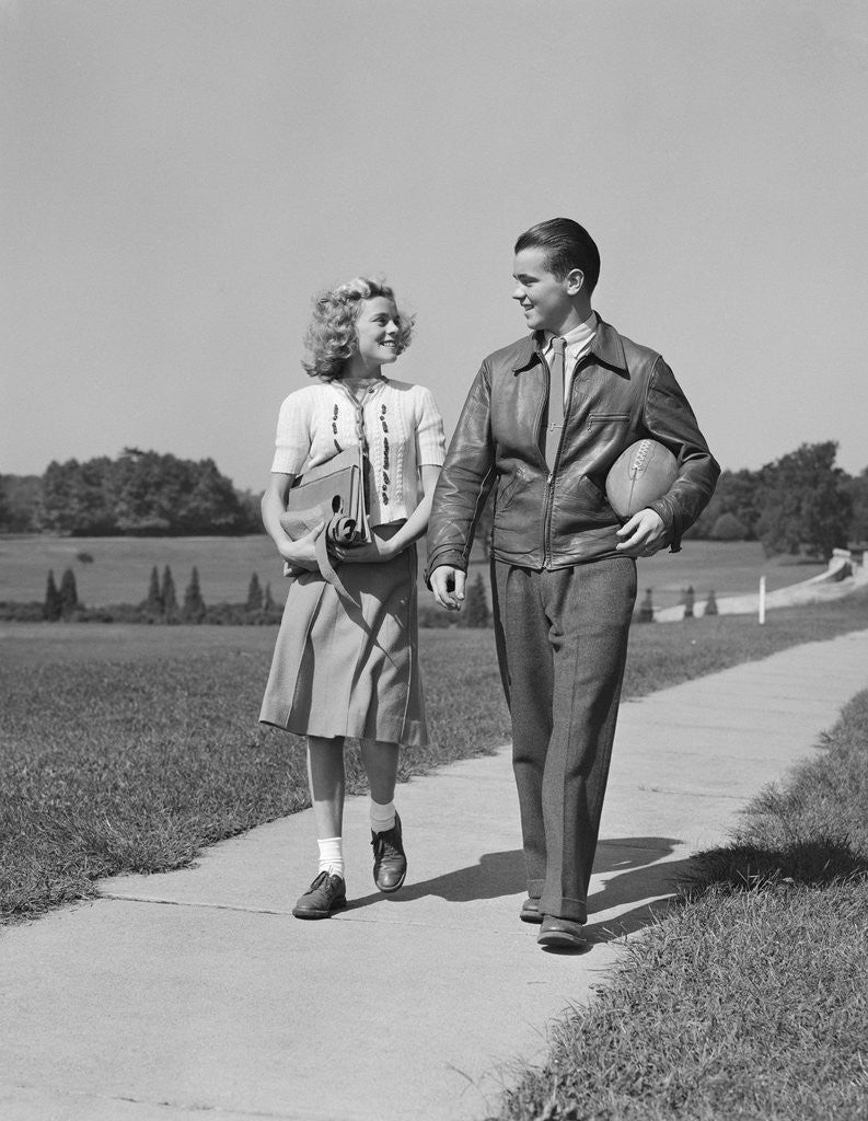 Detail of Teen couple walking down sidewalk girl with school books boy holding football by Corbis