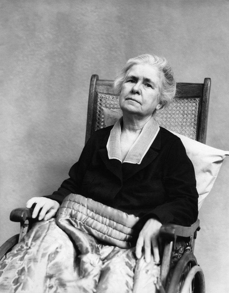 Detail of 1930s geriatric sad senior old woman in wheelchair looking at camera crippled by age with comforter blanket over legs by Corbis