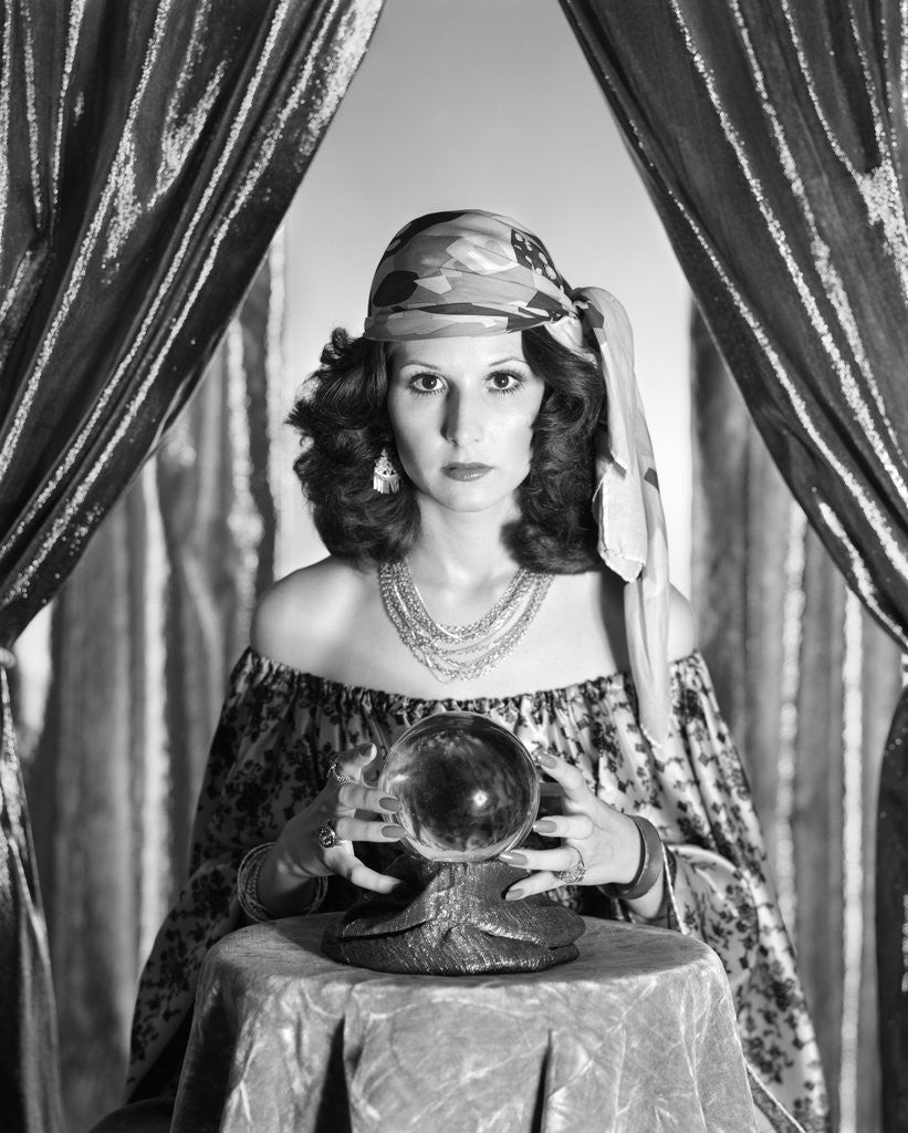 Detail of 1970s gypsy woman between sparkly curtains with hands around crystal ball staring into camera by Corbis