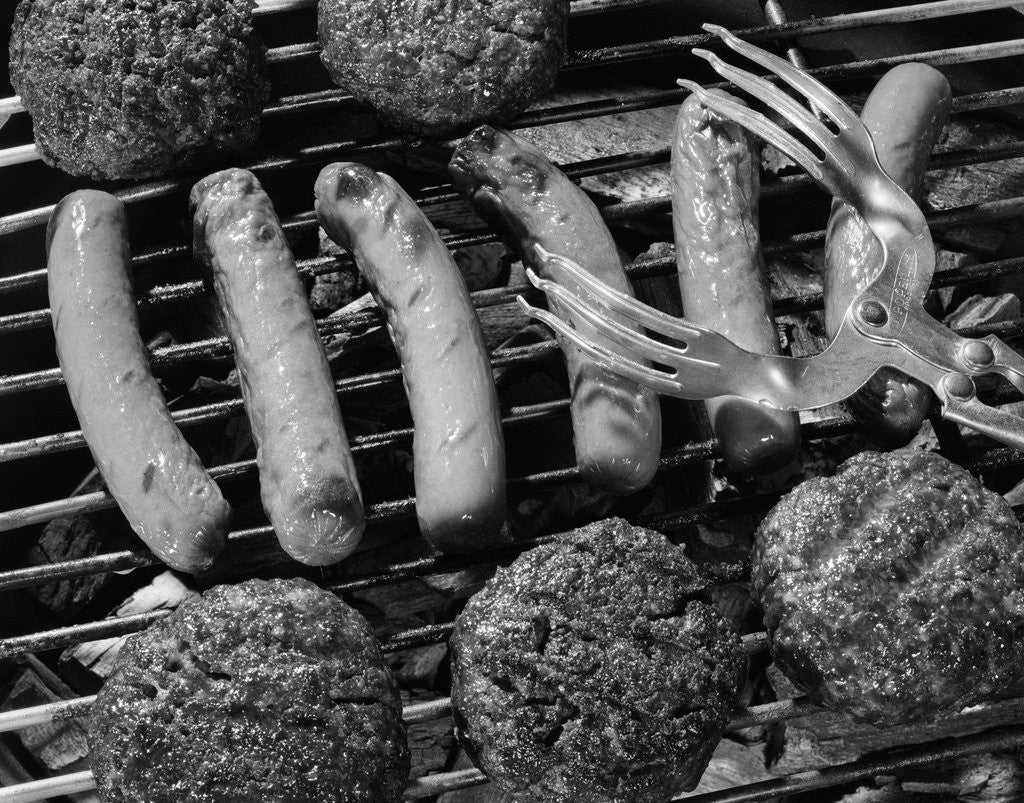 Detail of 1950s close-up of grill filled with hot dogs & hamburgers & tongs reaching down to turn them over by Corbis