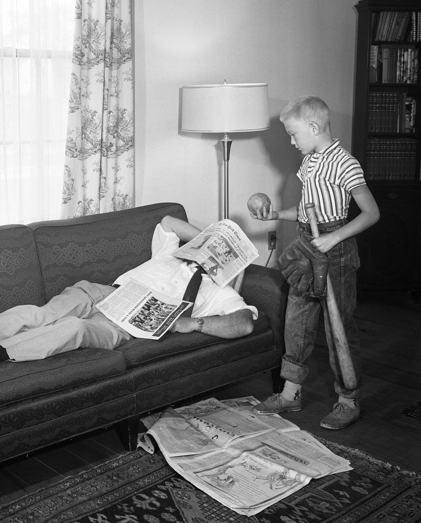 Detail of 1950s father lying on a sofa with newspaper over his head while son is standing over him with bat ball and baseball glove by Corbis