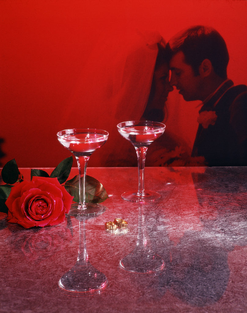 Detail of 1960s champagne flutes with rose and wedding rings in red filter with couple man woman bride groom in background by Corbis