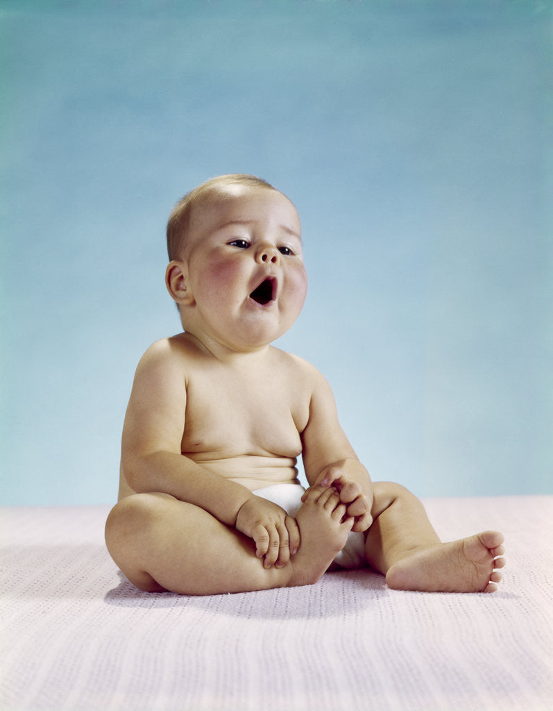 Detail of 1960s tired baby sitting holding on to one foot yawning facial expression by Corbis