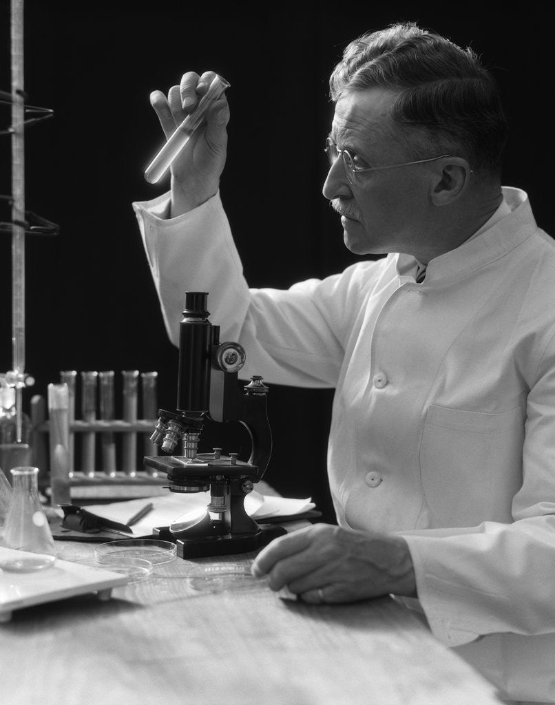 Detail of 1920s 1930s 1940s scientist lab technician in white coat looking at test-tube in front of microscope by Corbis